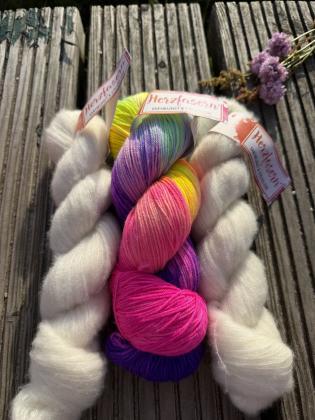 Handgefärbtes Garnset „Farbmagie“ von Herzfasern auf einer verwitterten Holzbank im Sonnenlicht fotografiert. In der Mitte liegt ein farbenfroher Strang Herzfasern No. 11 „Jolly“ mit Pink, Violett, Gelb und Türkis. Daneben liegen zwei fluffige Stränge Her
